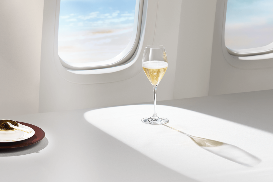 A glass of champagne set in front of an aircraft window in clear daylight, casting a shadow onto the ledge. On the left is a container of cavier is served on a plate.